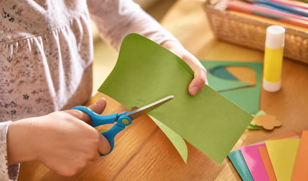 Feinmotorik fördern durch Basteln: Kinderhände schneiden präzise buntes Papier mit einer Kinderschere.