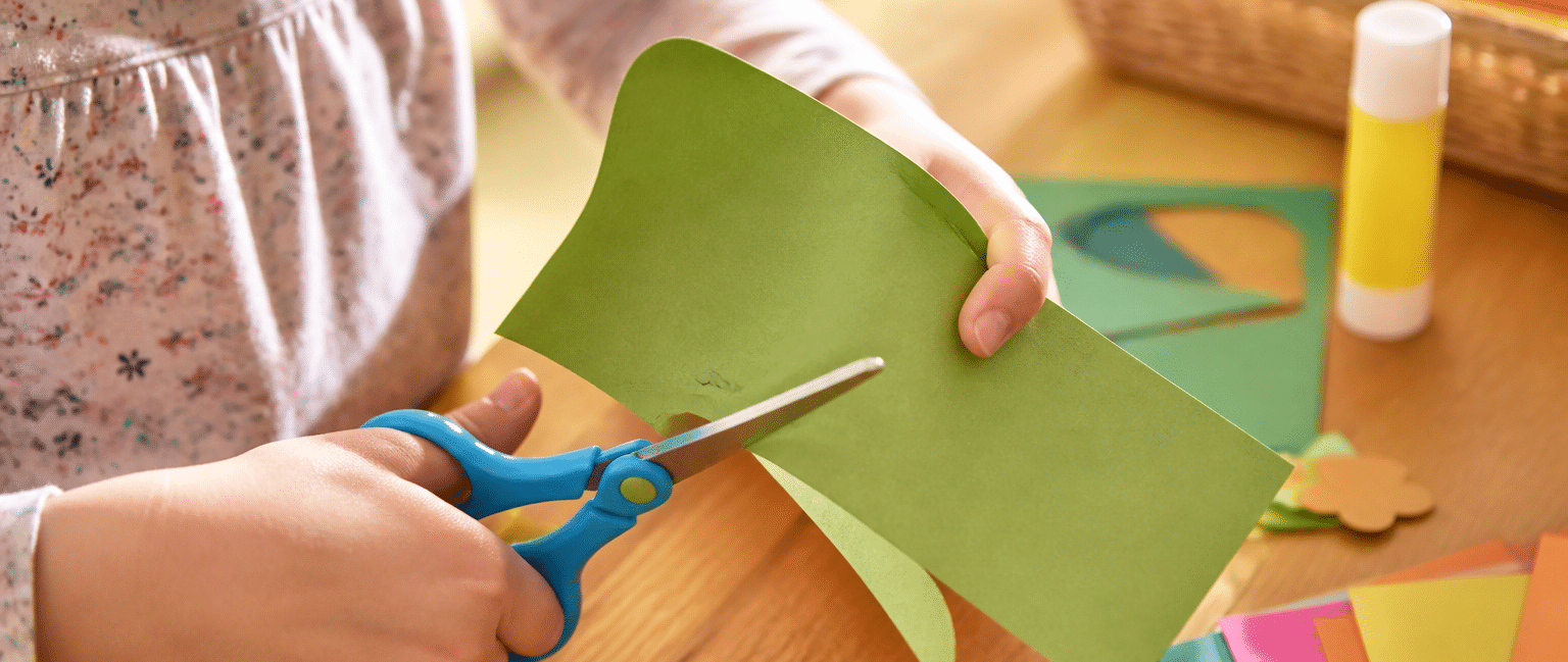Feinmotorik fördern durch Basteln: Kinderhände schneiden präzise buntes Papier mit einer Kinderschere.