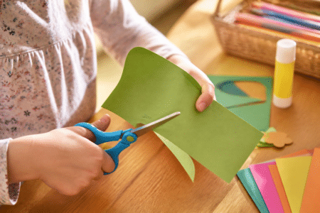 Feinmotorik fördern durch Basteln: Kinderhände schneiden präzise buntes Papier mit einer Kinderschere.
